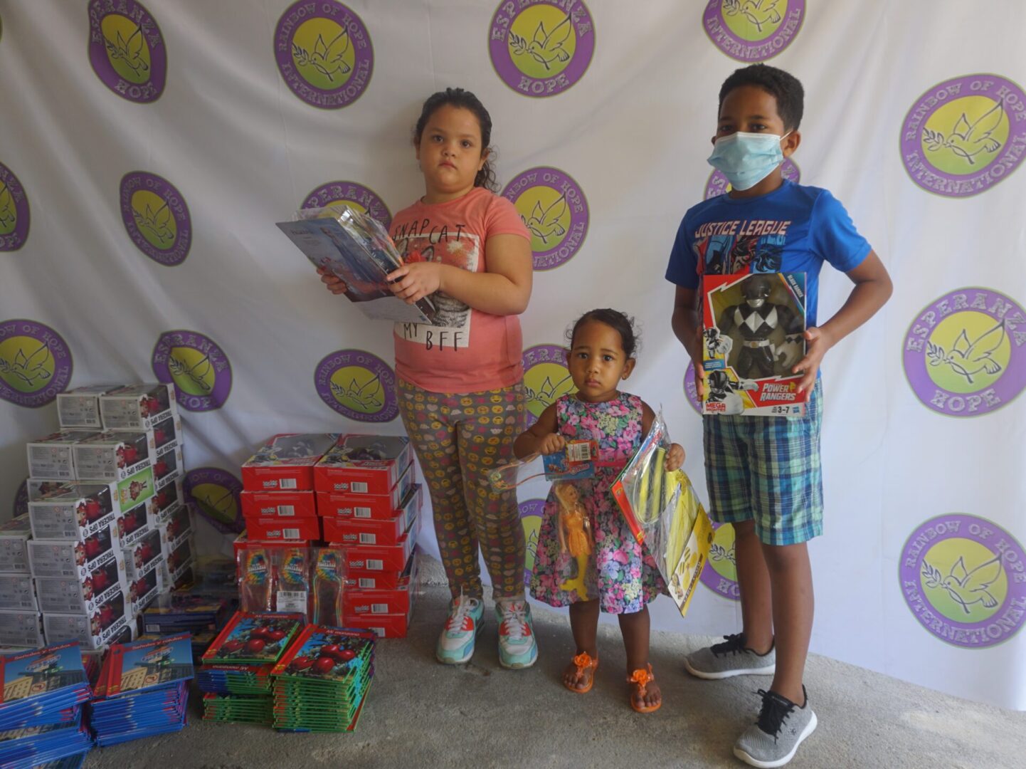Three children standing against the Esperanza-Hope backdrop with their toys and books