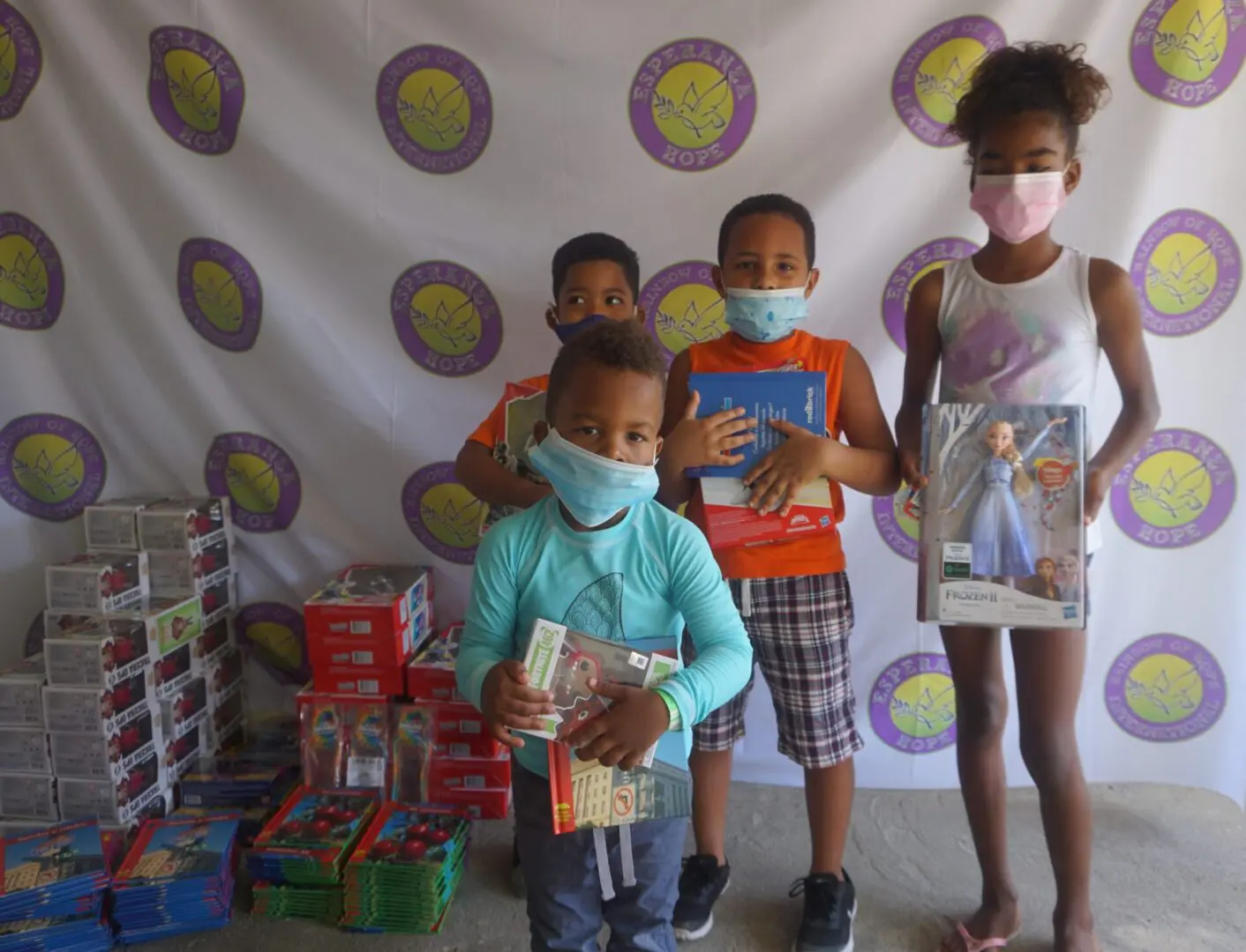 Four children standing against the Esperanza-Hope backdrop with their toys and books, batch 5
