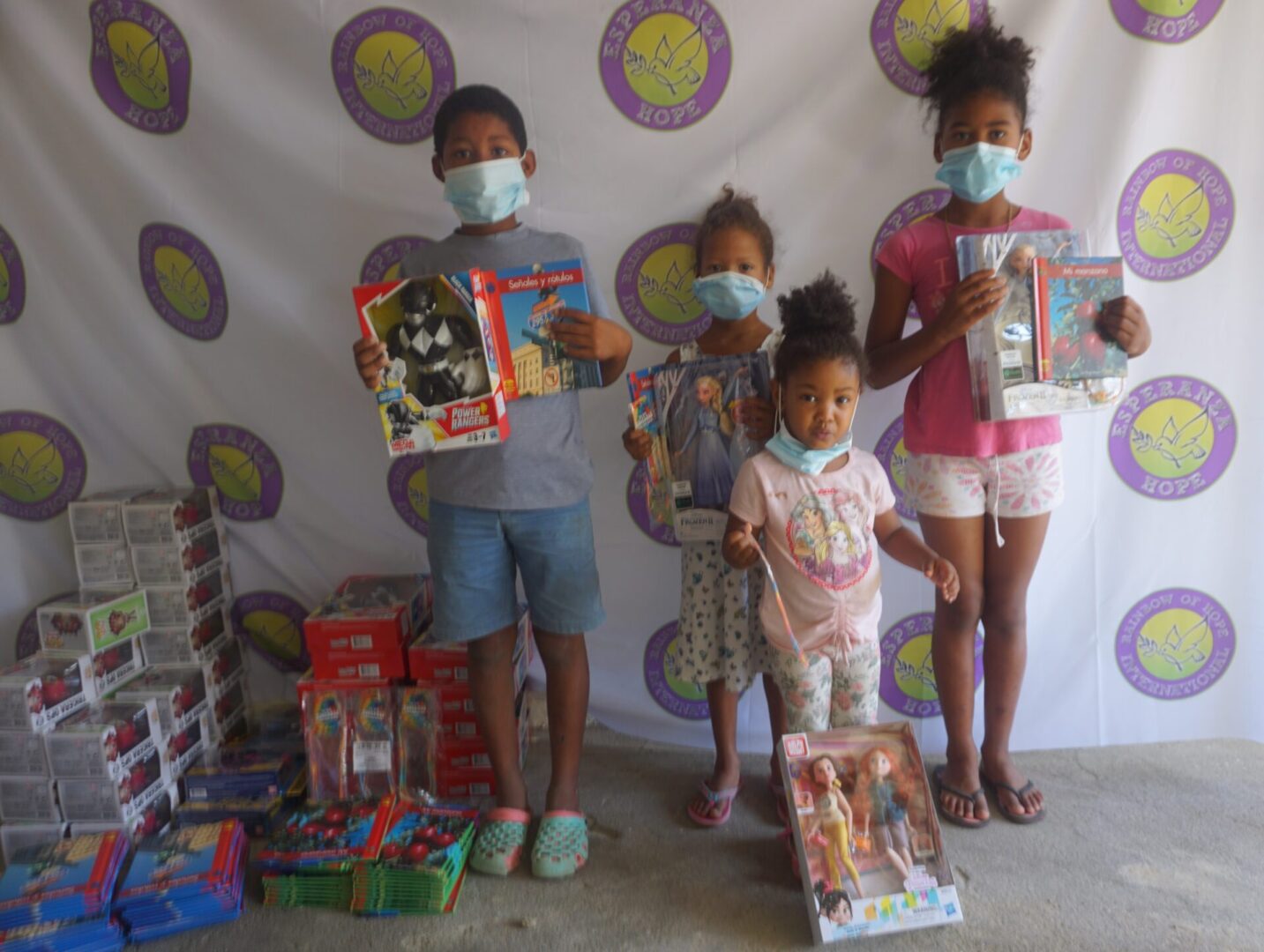 Four children standing against the Esperanza-Hope backdrop with their toys and books, batch 2