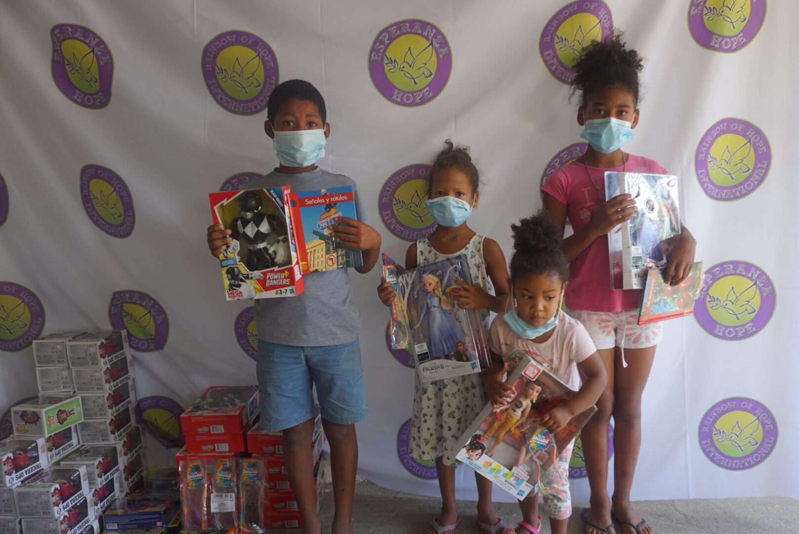 Four children standing against the Esperanza-Hope backdrop with their toys and books, batch 4