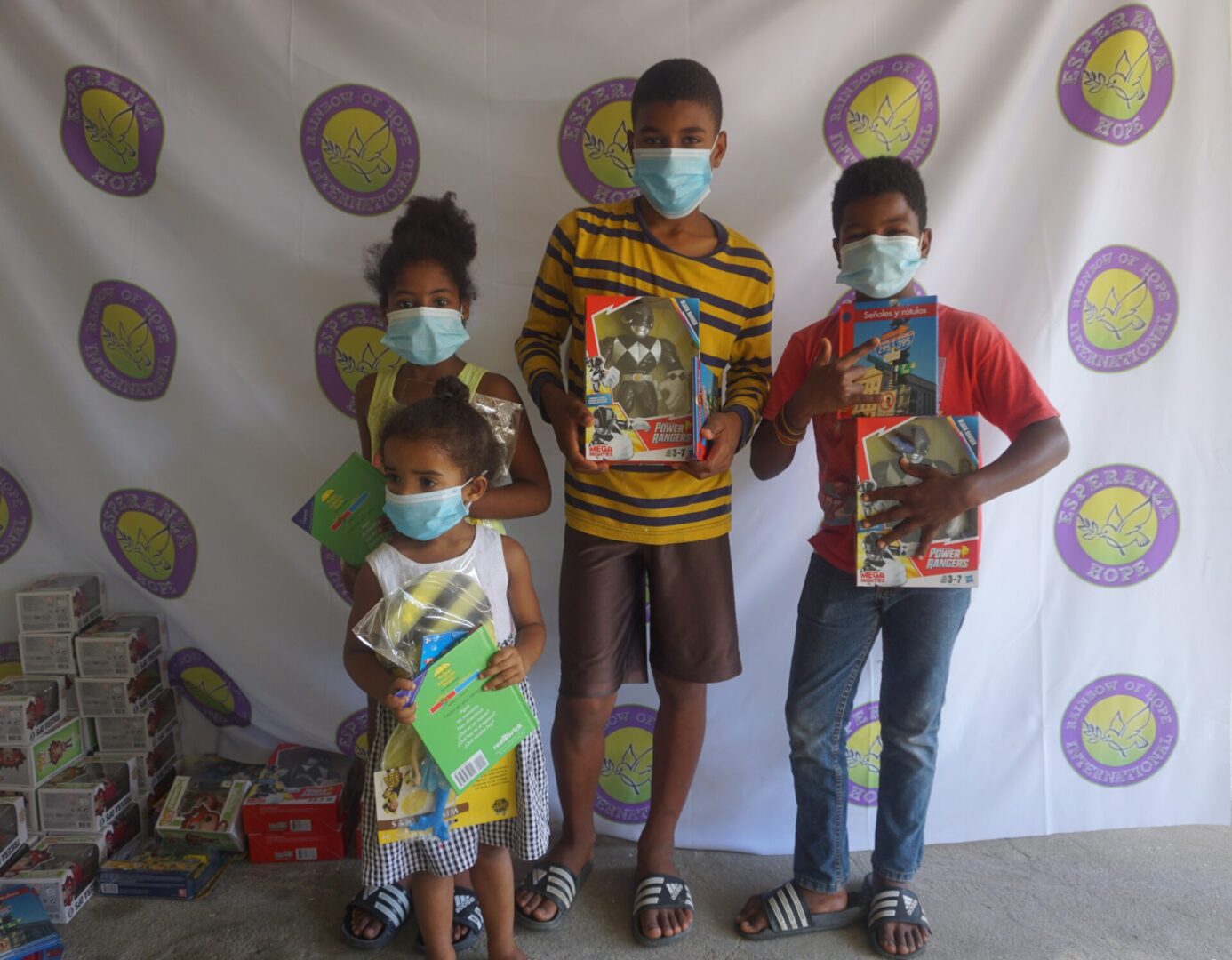 Four children standing against the Esperanza-Hope backdrop with their toys and books, batch 3