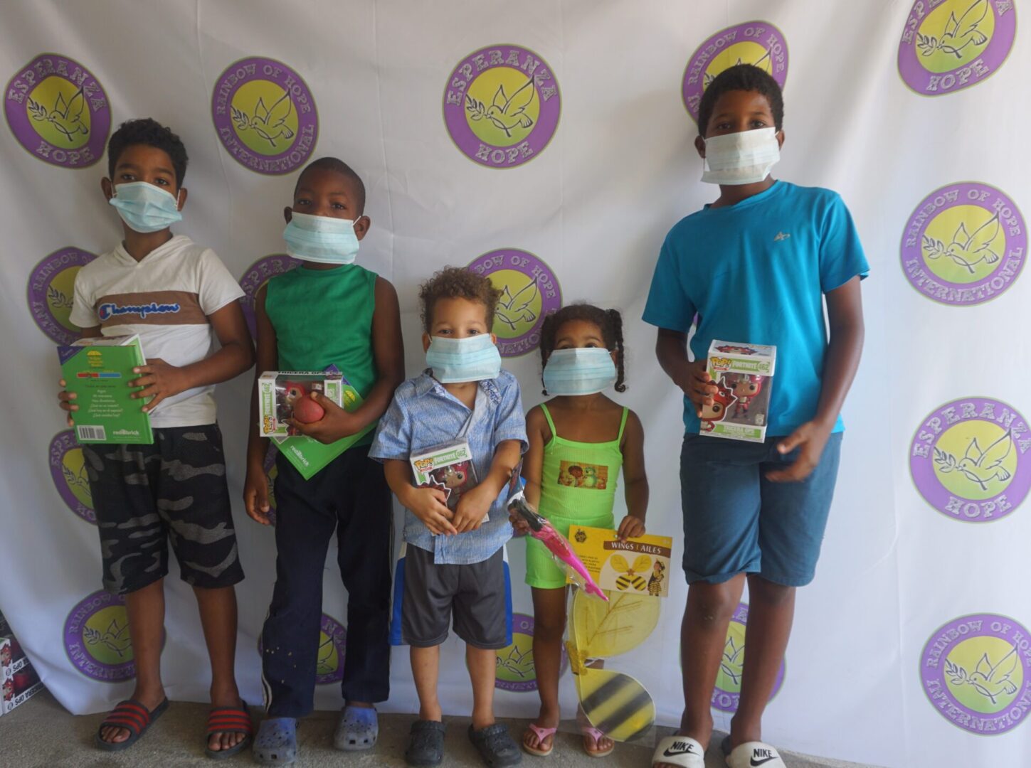 Five children standing against the Esperanza-Hope backdrop with their toys and books