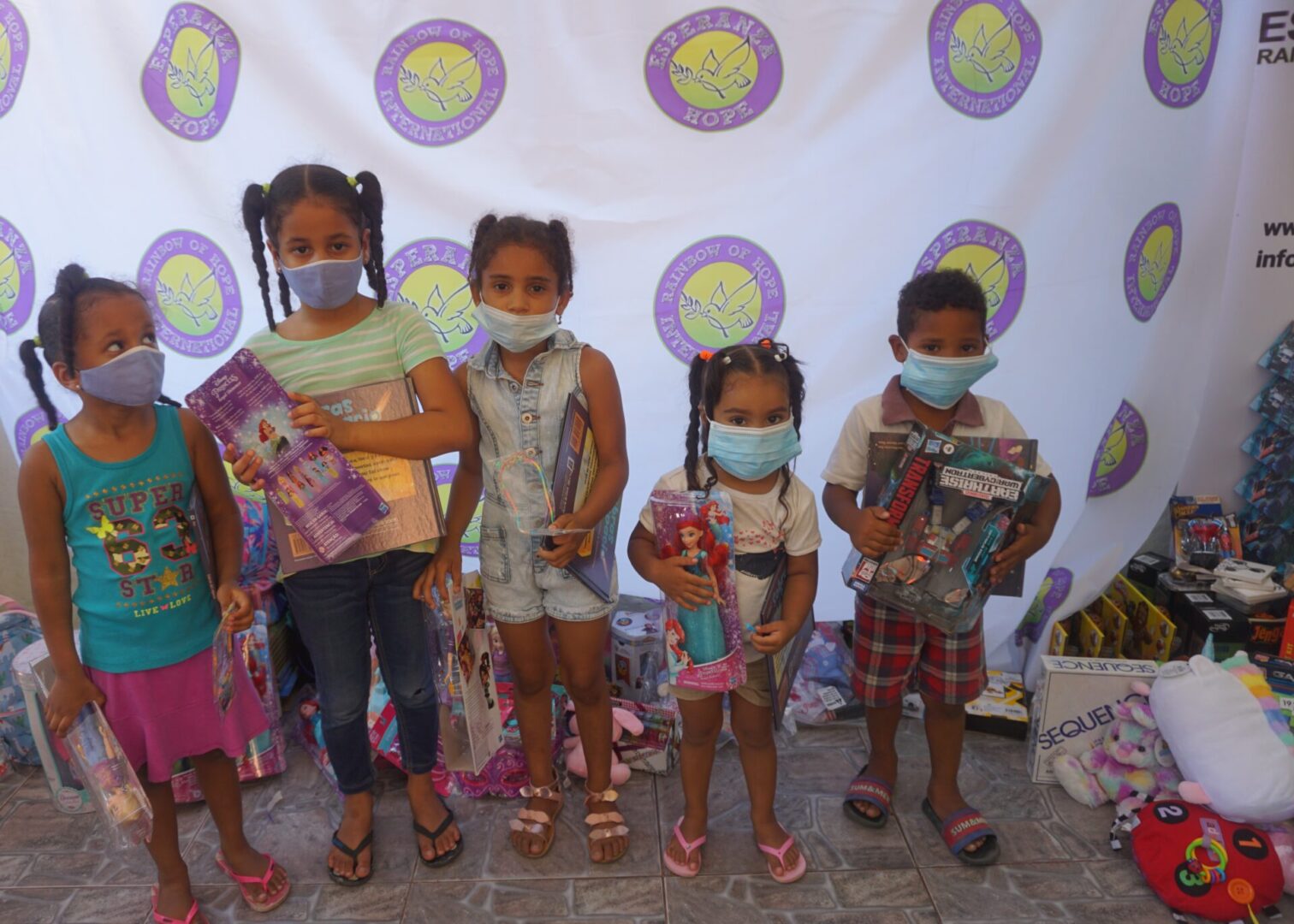 Five children standing against the Esperanza-Hope backdrop with their toys and books, batch 6
