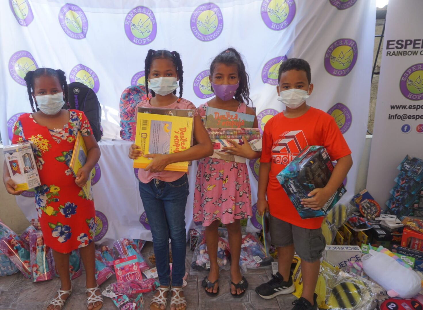 Four children standing against the Esperanza-Hope backdrop with their toys and books, batch 14
