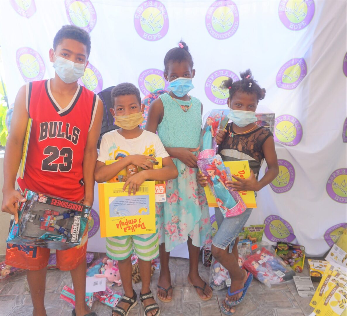 Four children standing against the Esperanza-Hope backdrop with their toys and books, batch 13