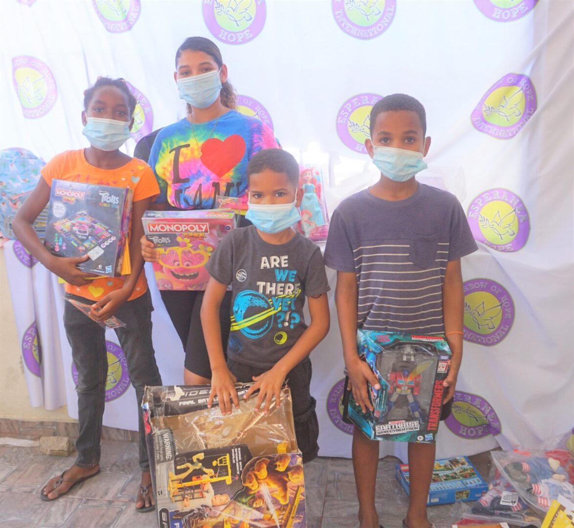 Four children standing against the Esperanza-Hope backdrop with their toys and books, batch 11