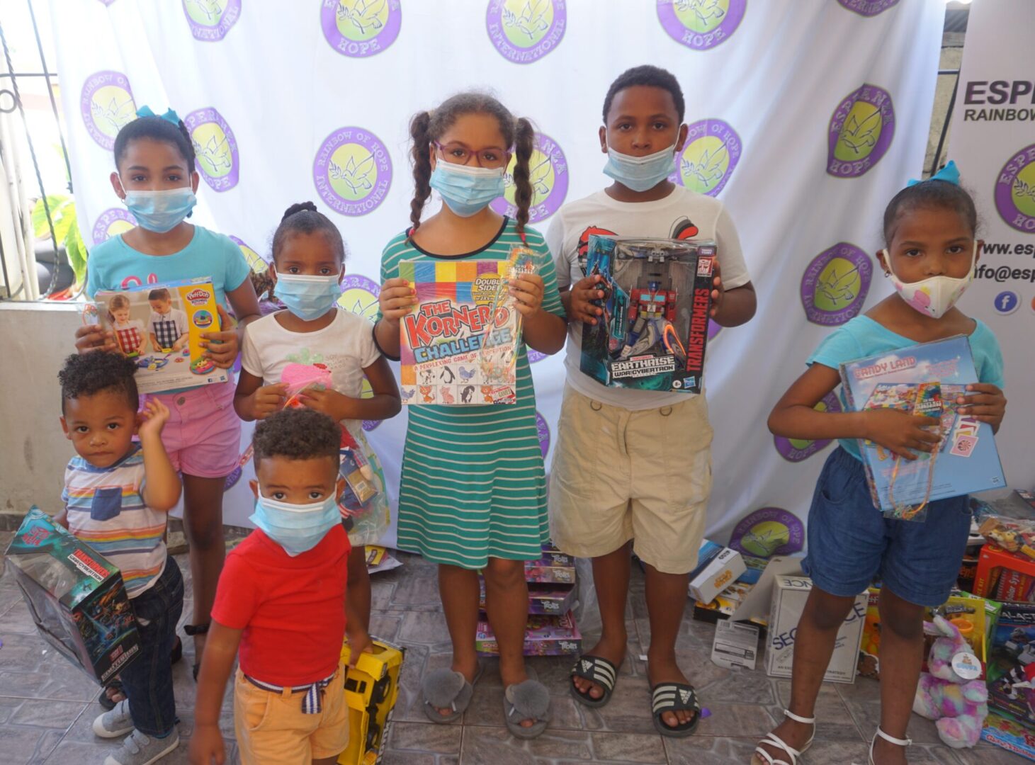 Seven children standing against the Esperanza-Hope backdrop with their toys and books