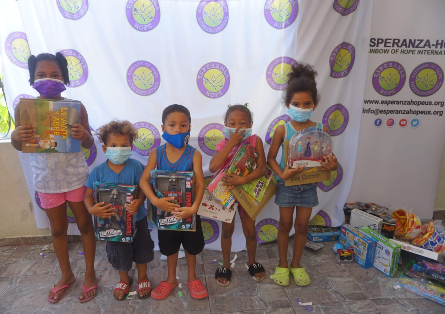 Five children standing against the Esperanza-Hope backdrop with their toys and books, batch 8