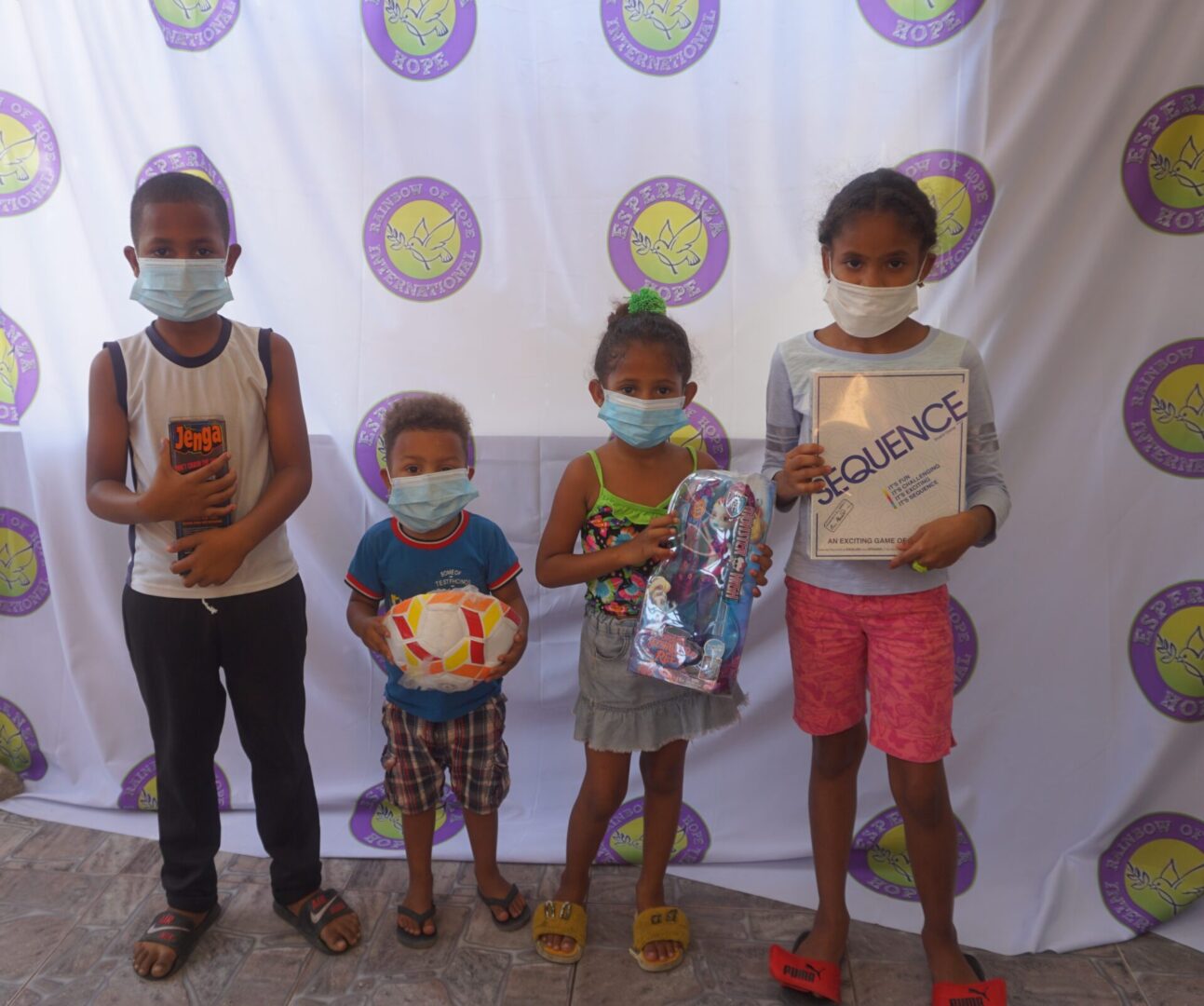 Four children standing against the Esperanza-Hope backdrop with their toys and books, batch 12