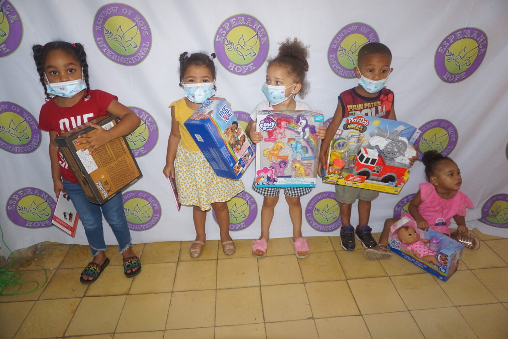 Five children standing against the Esperanza-Hope backdrop with their toys and books, batch 10